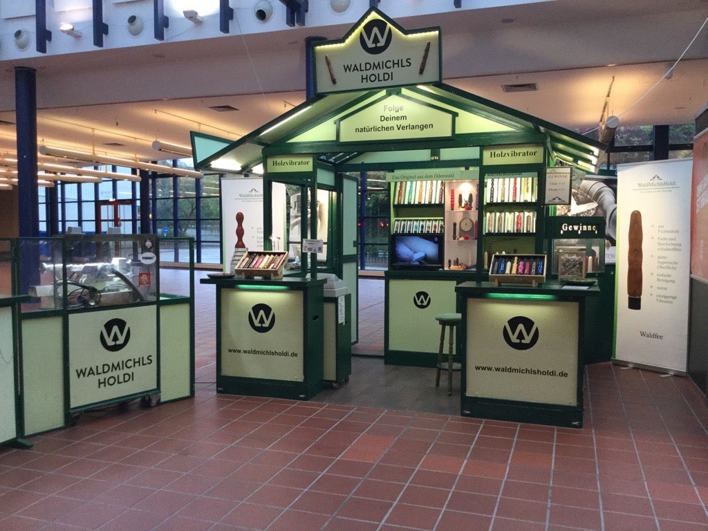 Eine grüne Holzhütte steht in einem Einkaufszentrum als Verkaufsstand daneben eine Drechselbank. Im Regal in der Hütte liegen Holzdildos und Holzvibratoren in verschiedenen Farben aus.